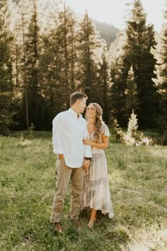 a man and woman are standing in the grass near some trees with their arms around each other