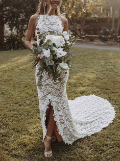 a woman in a white lace dress holding a bridal bouquet standing on the grass