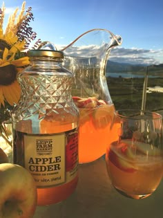 a pitcher of apple cider next to two glasses with apples and flowers in the background