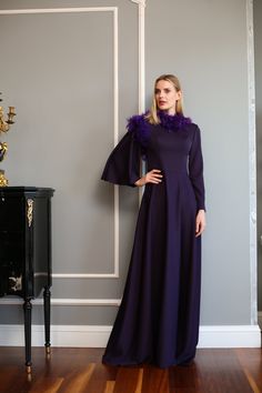 a woman in a long purple dress standing next to a black cabinet and gold vase