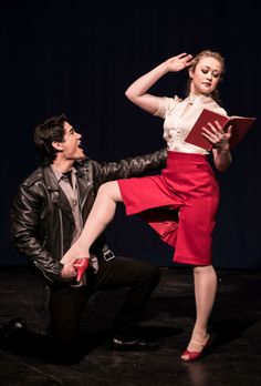 a man kneeling down next to a woman in a red skirt and white shirt holding an open book