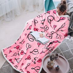 a woman wrapped in a pink heart print blanket reading a book while sitting on a chair