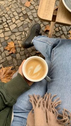 a person holding a cup of coffee while sitting on a bench with their legs crossed