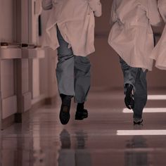 two men in white lab coats walk down a hallway together, both wearing black shoes