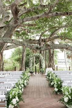 an outdoor ceremony setup with white chairs and greenery on the aisle, surrounded by trees