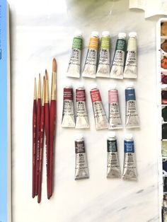 an assortment of paints and brushes on a table with watercolors in the background