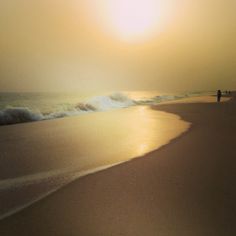 two people are walking on the beach near the ocean as the sun goes down in the distance