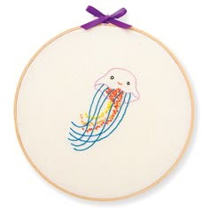 a cross - stitch jellyfish with purple ribbon hanging on a white wall mounted hoop