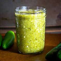 a glass jar filled with green stuff next to three green peppers