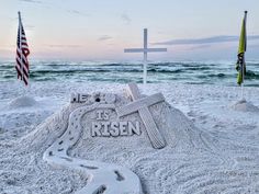 there is a sand sculpture that says it's risen on the beach with an american flag in the background