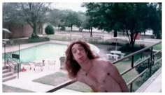 a man with no shirt standing in front of a swimming pool and looking at the camera