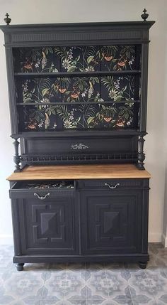 an old black china cabinet with floral wallpaper on the top and drawers below it