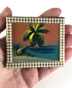 a hand holding a miniature painting of a palm tree on the beach with pearls around it