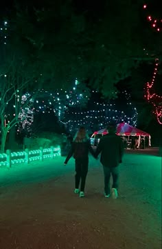 two people walking down a path with christmas lights on the trees and behind them are benches