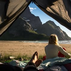 a woman sitting on top of a bed under a tent next to the ocean with mountains in the background