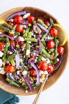 a wooden bowl filled with salad and topped with tomatoes, onions, asparagus, red onion, olives, feta cheese and lemon wedge