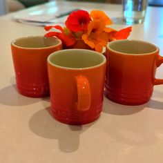 three orange mugs sitting on top of a white table next to a vase with flowers