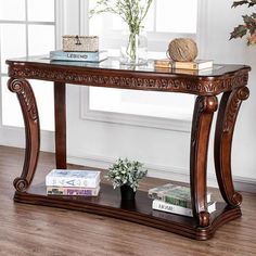 a wooden console table with glass top and shelves on the bottom, in front of a window