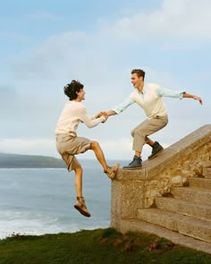 two men are jumping off the edge of a stone wall and one is holding onto another man's hand