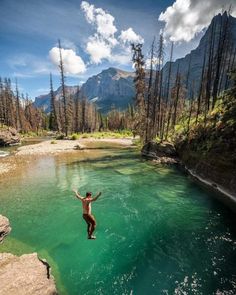 a man is jumping into the water from a cliff in front of some trees and mountains