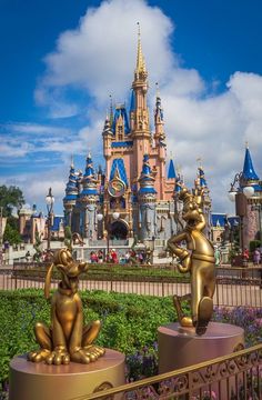 two gold statues in front of a castle with blue sky and clouds behind them at disney world