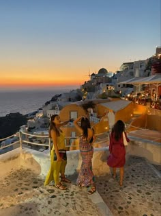 three women standing on the edge of a cliff looking at the ocean and buildings in the background