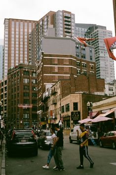 people crossing the street in front of tall buildings with kites flying above them on a cloudy day