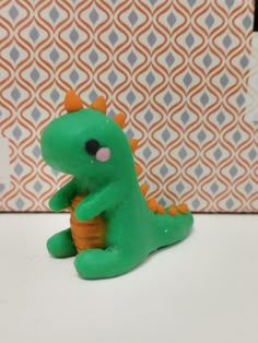 a green toy dinosaur sitting on top of a white table next to a patterned wall