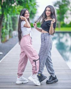 two young women standing next to each other on a wooden walkway near a pool and trees