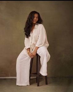 a woman sitting on top of a wooden chair wearing white clothing and gold bracelets