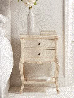 a white vase with flowers sitting on top of a small table next to a bed