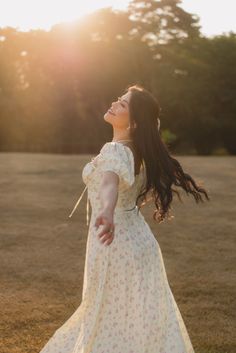 a woman in a white dress standing on top of a grass covered field with the sun behind her