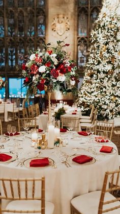 a table set up with white linens, red napkins and gold chairs in front of a christmas tree