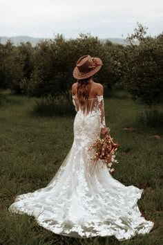 a woman in a wedding dress and hat is standing in the grass with her back to the camera