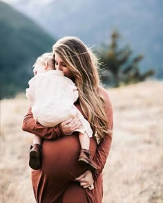 a woman holding a baby in her arms while standing on top of a grass covered field