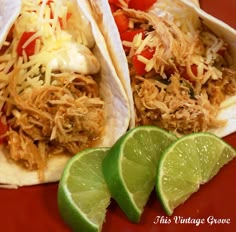 two tacos with shredded chicken, tomatoes and cheese on them next to lime wedges