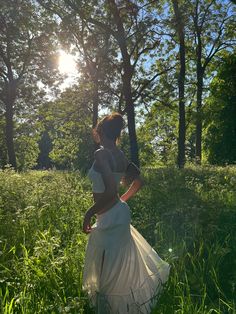 a woman in a white dress is walking through the grass with trees and sunlight behind her