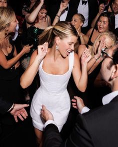 a woman in a white dress surrounded by other people at a party with her hands up to her face