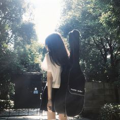 a woman holding a guitar in her right hand and looking at the ground with trees behind her