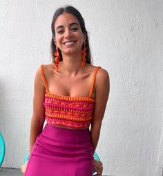 a woman in a purple skirt and orange top smiles at the camera while leaning against a wall