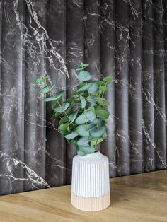 a white vase filled with green leaves on top of a wooden table next to a marble wall