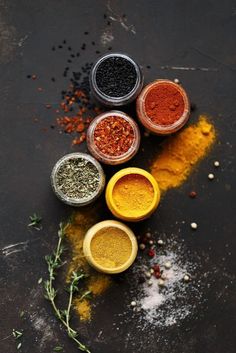 five different colored spices in small bowls on a black surface with sprigs of rosemary