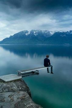 a man sitting on a dock in the middle of a body of water with mountains in the background