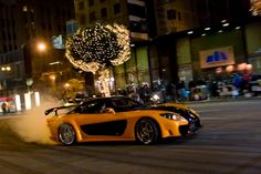 a yellow sports car driving down a street at night with lights on the buildings in the background