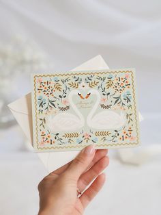 a person holding up a card with two swans on it and flowers around the edges