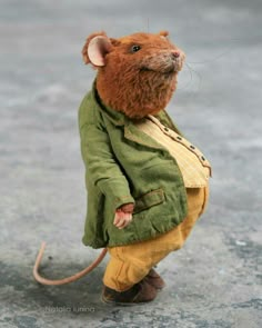 a stuffed mouse in a green jacket and yellow pants is standing on the pavement with its head turned to the side