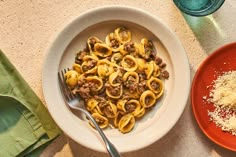 a bowl filled with pasta and meat next to a red plate full of grated parmesan cheese