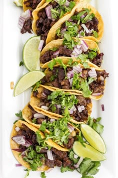 three tacos on a white plate with limes and cilantro wedges