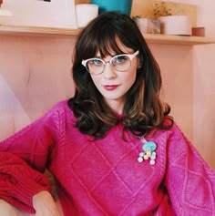 a woman with glasses sitting in a chair wearing a pink sweater and flower brooch