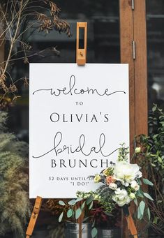 a welcome sign with flowers and greenery is displayed in front of a window for an outdoor bridal brunch